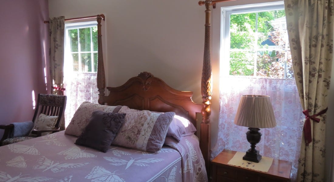 Four poster bed near two sunny windows with side table and lamp