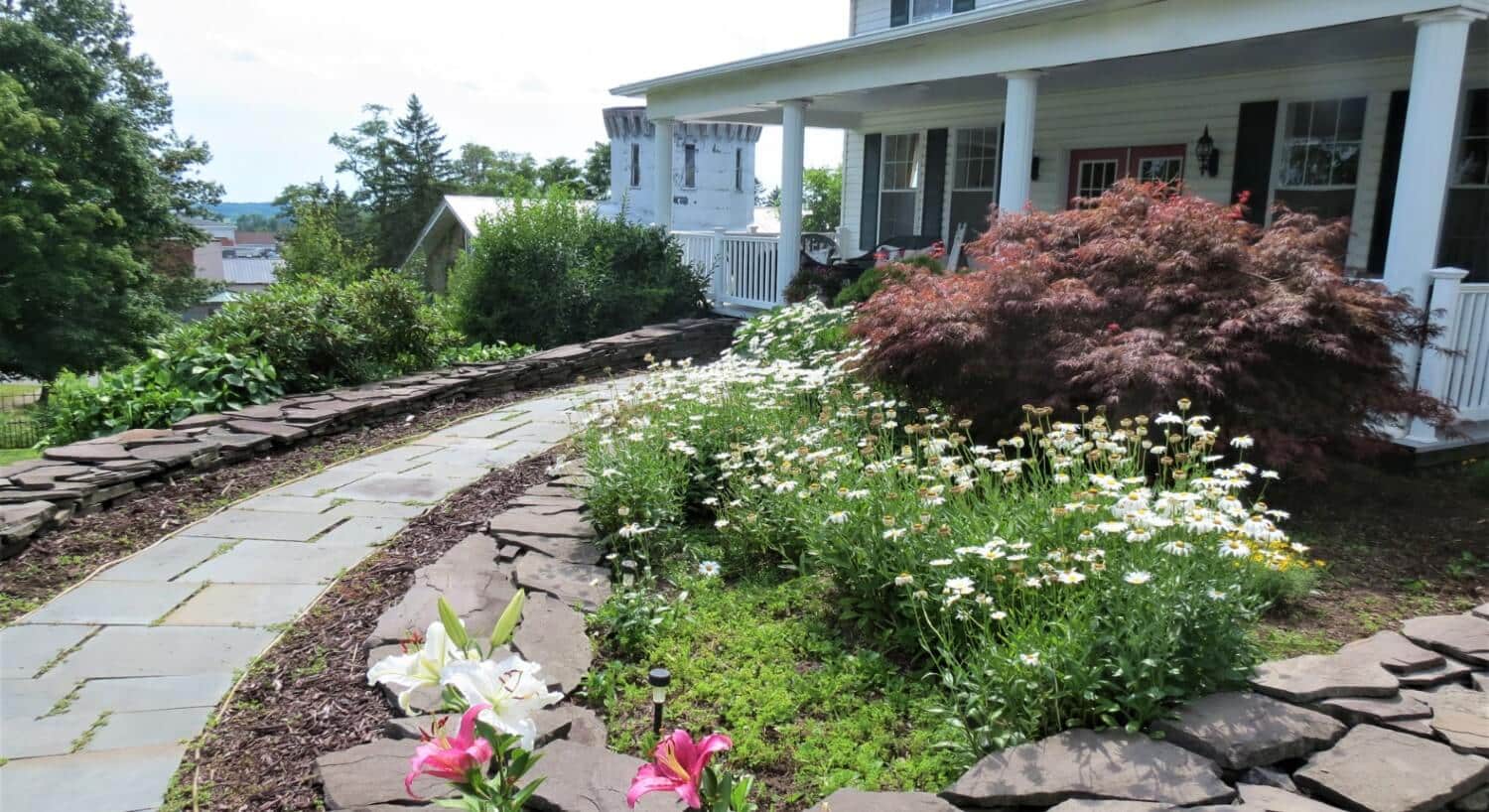 Front porch of a white home with red and green bushes and a curved stone path leading from front door