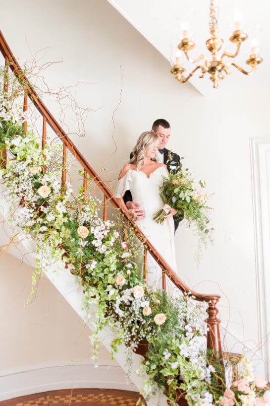 Blonde bride and brunette groom in gown and tux stand on a staircase decorated with pink and white flowers. 