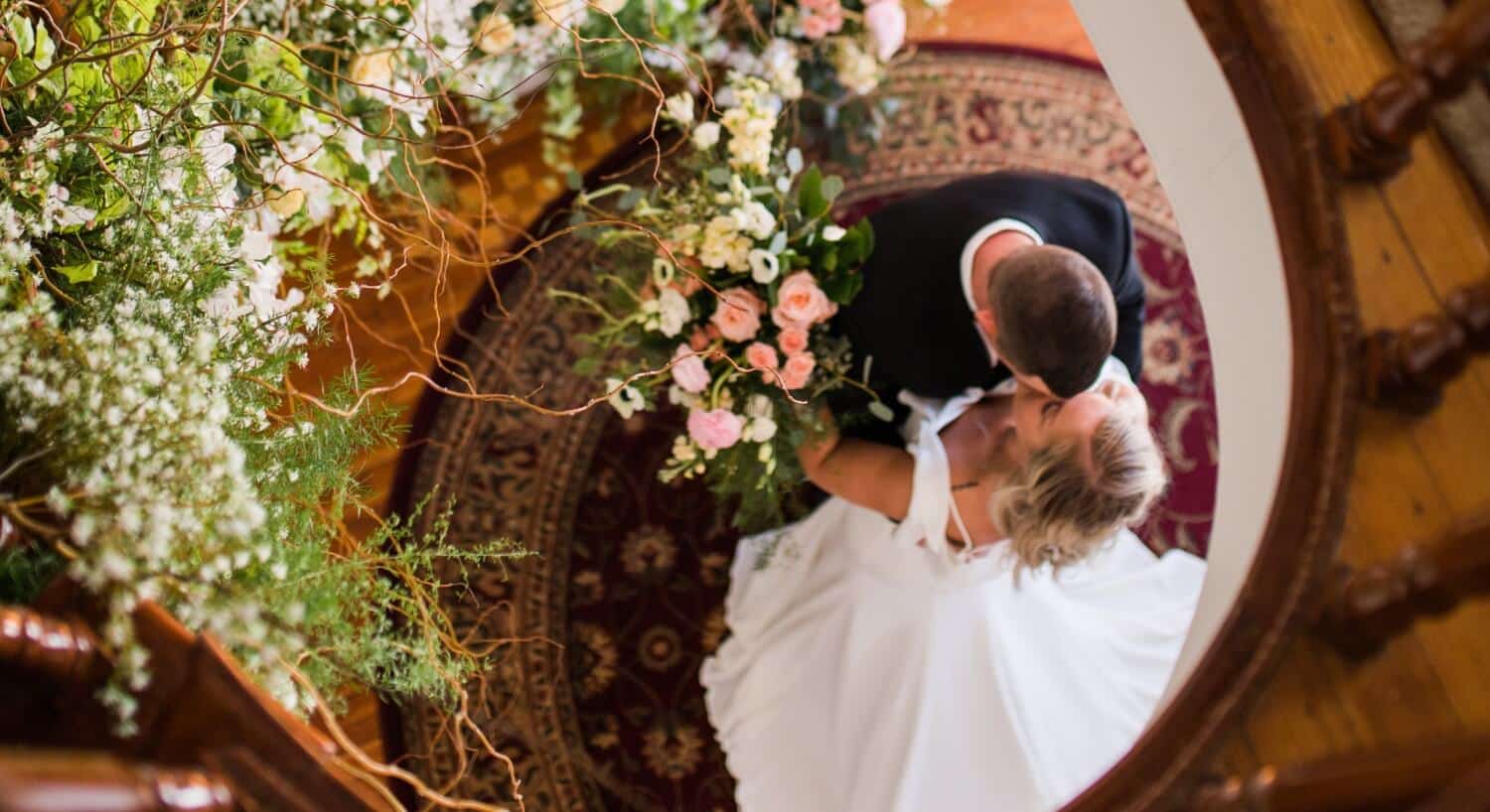 Bride and groom embracing at the bottom of a lavish brown spiral staircase covered in flowers