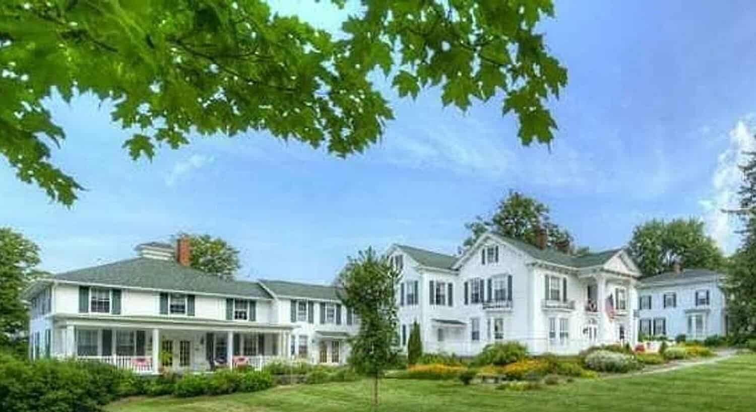 Expanisve white mansion with green roof and shutters surrounded by green lawn and trees. 