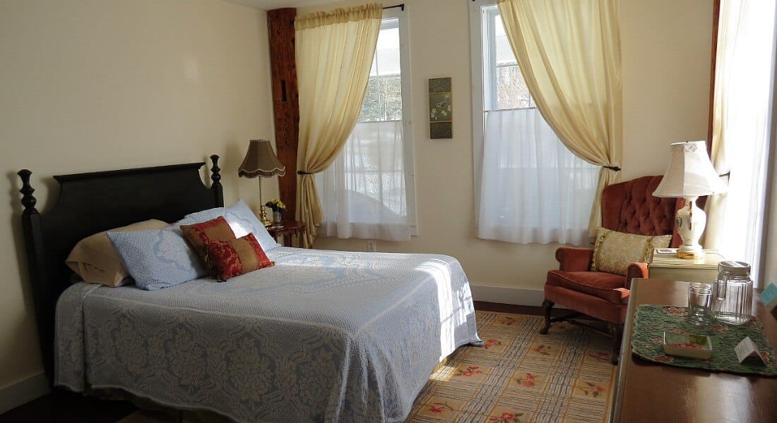 Bedroom made light by several large windows holds a wooden queen bed and chair with a dresser.