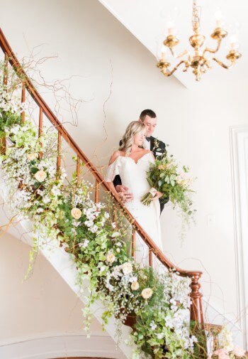 Blonde bride and brunette groom in gown and tux stand on a staircase decorated with pink and white flowers.